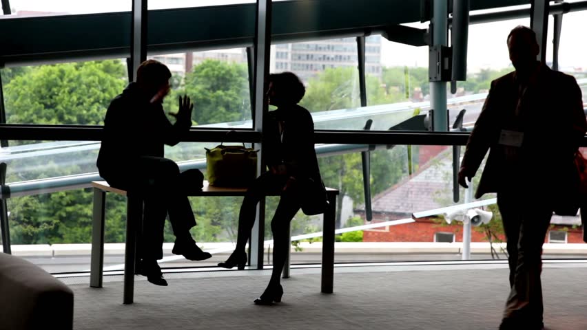 Man and a woman are sitting at the table near the window, two man are passing past them.