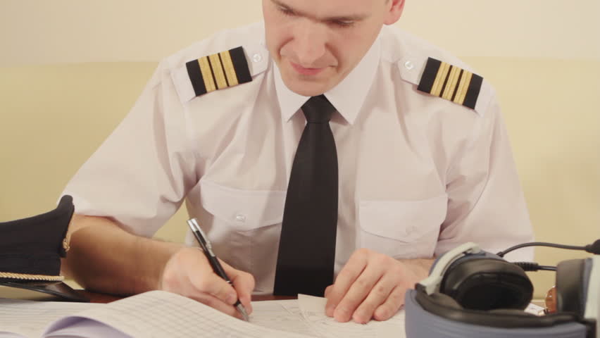 Airline pilot wearing hat, shirt with epaulets and tie filling in and checking papers flight plan, log book and weather forecast. Headset on the table