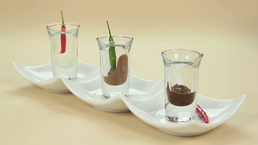 A chili being dipped into chocolate in a shot glass in a row of glasses.