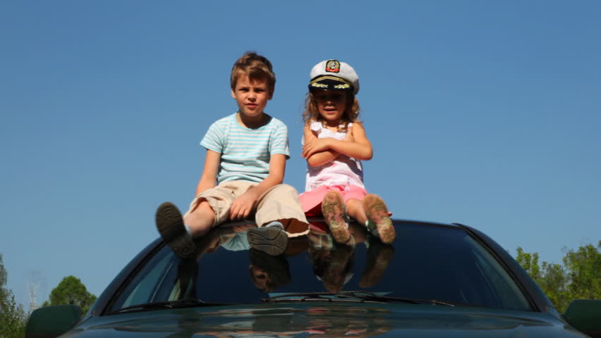 boy and little girl in captain's cap sit on roof of car, cheerfully waving their hands, then he taking off her hat puts it on himself against blue sky summer