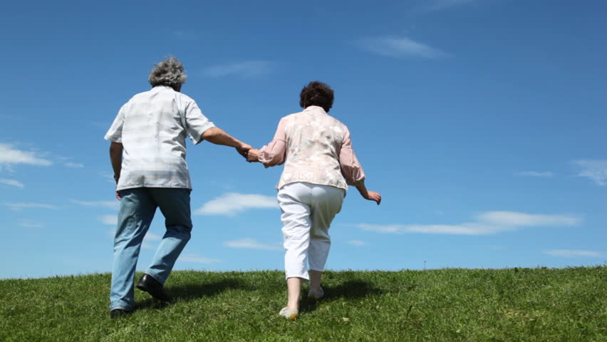 mature couple leaves hand in hands through grassy hill against sky summer