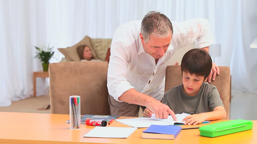 Little dark-haired boy doing homework after school in the living room with the help of his grandfather 