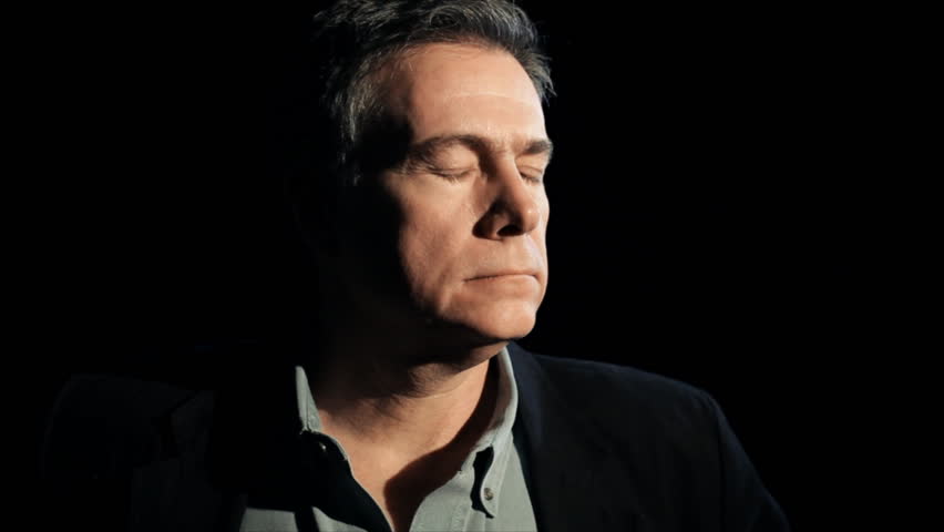A dark backdrop and harsh lighting frames a mature melancholy man stopping and staring solemnly into the camera.