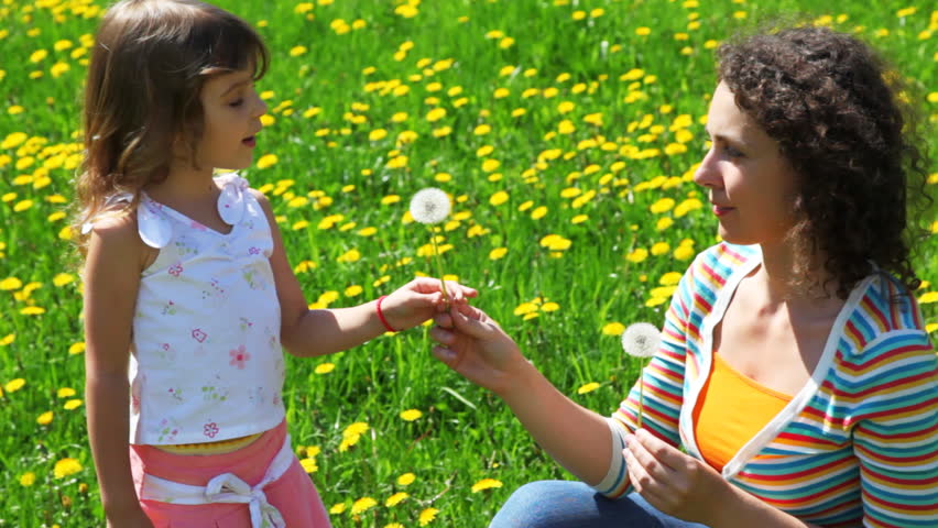 little daughter playfully blows off dandelion in face of mother on green lawn spring day