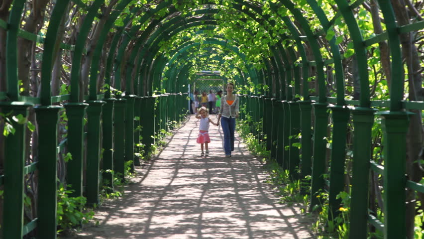 mother and her little daughter go through arched corridor braided green plants by spring day