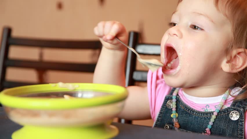 Baby girl is eating porridge from a green plate and moms arm wipes her mouth