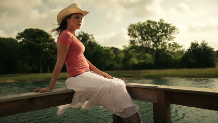 A pretty girl in a long white skirt wearing a cowboy hat sits on the railing of a small pier built on a large pond.