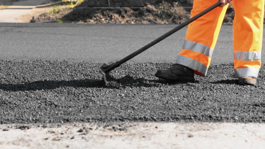 Road construction worker leveling asphalt using rake while in the back road roller passing by