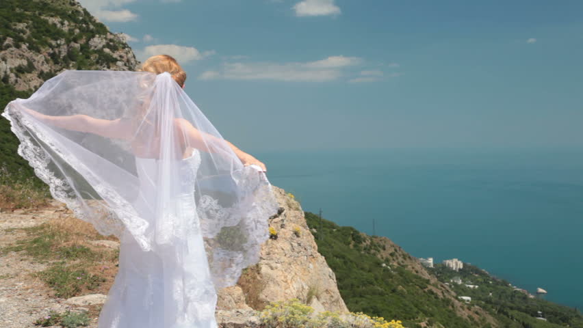bride stands on cliff with a flowing veil on the wind against the blue sea and sky
