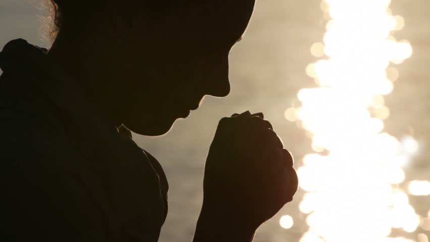 Silhouette of woman head with sunshine reflected in water behind, holding two fists together and bowed head, then rises head up, after bowed head again