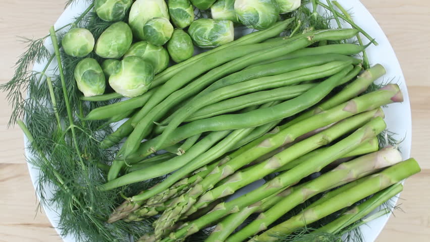 Vegetable platter of asparagus, green beans, dill sprigs and Brussel sprouts rotate on white plate against wooden kitchen counter background. 1080p