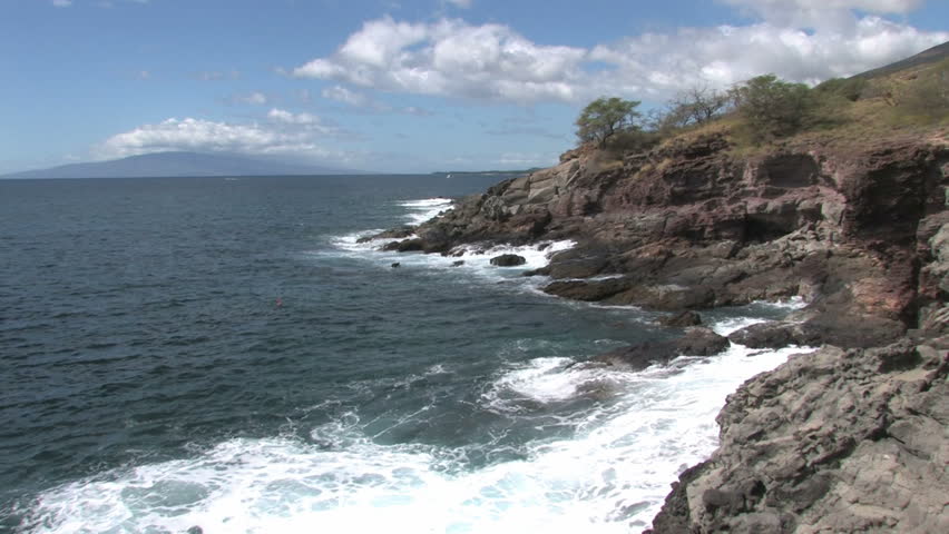Rocky shoreline on the island of Maui in Hawaii. A man swimming and spear fishing in the rough water. Beautiful water with cliff overlooking water. Island of Lanai on horizon.