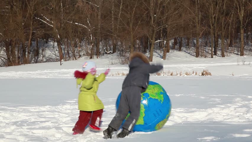 Two kids boy and girl play with inflated ball with earth map texture, they push it and jump on it when fell on snow, at sunny winter day