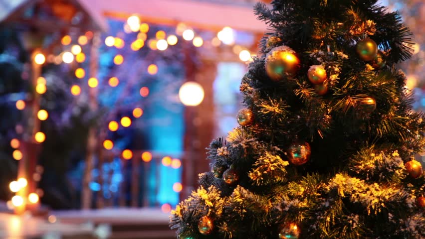 Christmas tree with balls stand in front of blurred wooden house decorated by garland