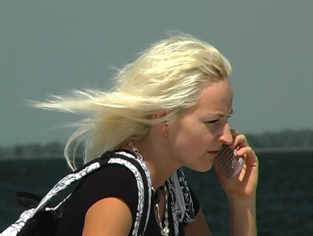 Close-up of a beautiful blonde talking on her cell phone outdoors at a lakefront on a bright, sunny, and breezy day. She faces into the wind to talk.  Ends with a freeze frame.