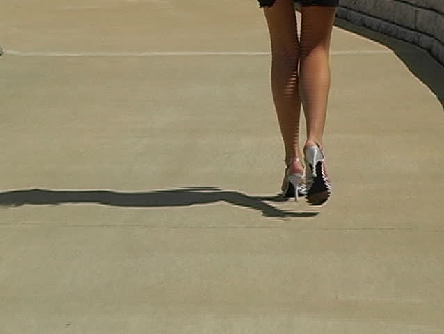 Close-up shot of a pair of beautiful female legs wearing high heels walking away from the camera. Zooms in to follow.