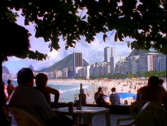 Silhouetted people sit at tables in an outdoor cafe with Copacabana beach in the background.