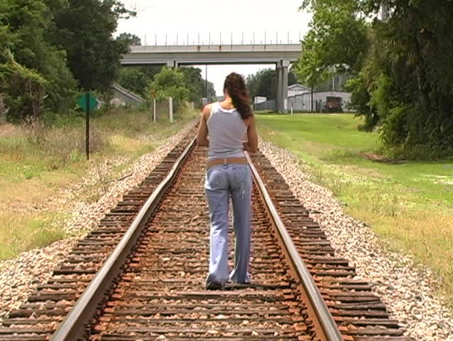 A beautiful Latina brunette walks away from the camera down a lonely stretch of railroad track.  Full length.  Camera zooms in to follow.