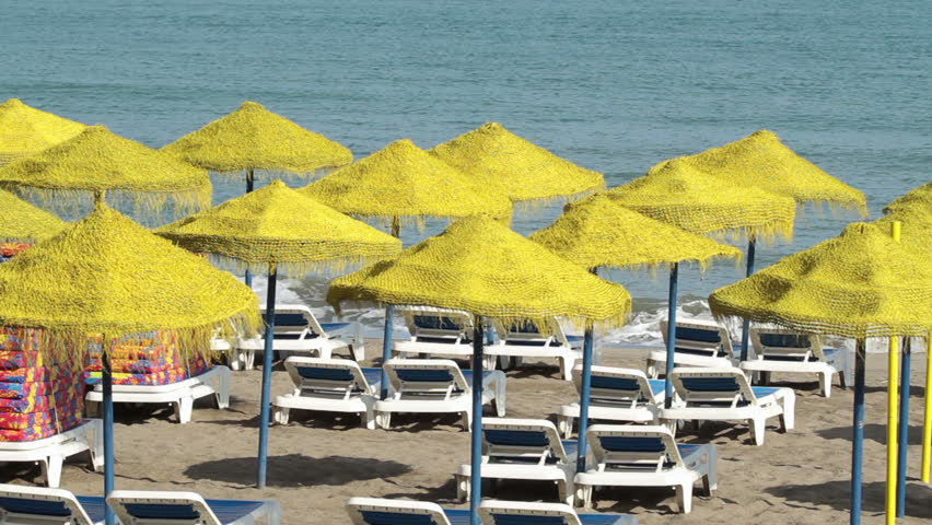 Beach umbrellas on beach near Malaga Spain on the shore of the Mediterranean Sea. Beautiful yellow shade and white lounge chairs against blue ocean water and sand.
