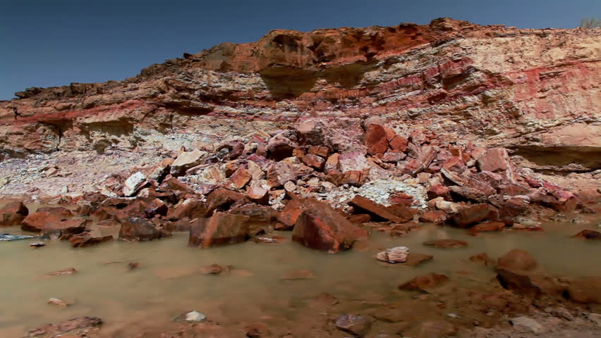 Dolly move left to right along a slow moving stream at the base of a rocky landslide from a visible cliff face.  This Image is from the desert floor of the Mitzpe Ramon Crater in Israel.