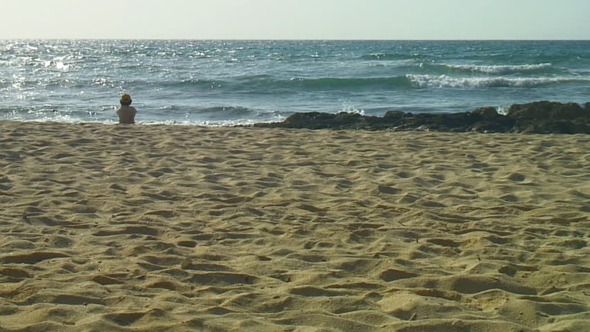 Woman sits alone on beach gazing at waves in morning sun.