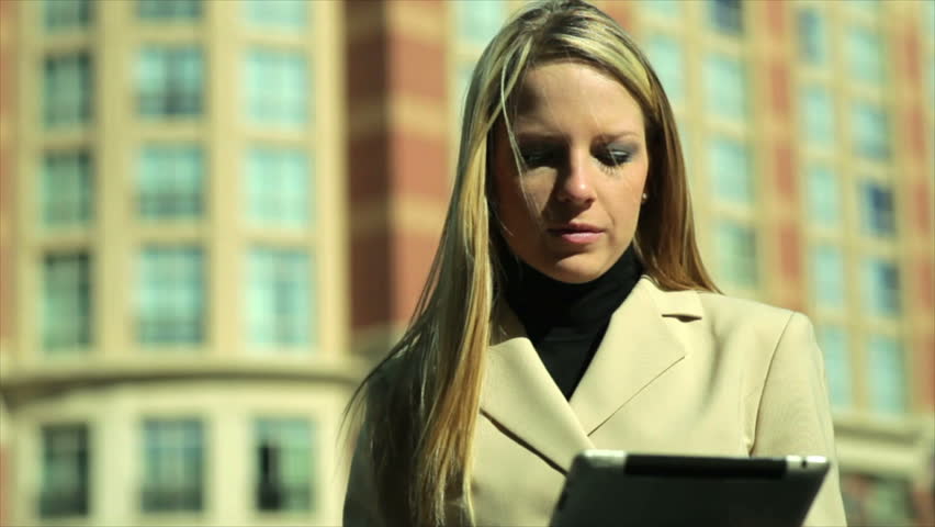 A pretty blond businesswoman in an urban setting on a bright breezy sunny day uses her digital or electronic tablet.