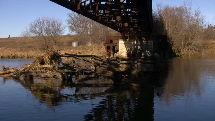 An old metal train bridge bridge crosses over the Grand River.  The river is gently flowing as the bridge is reflected. A tree is caught in the current. (High Definition)