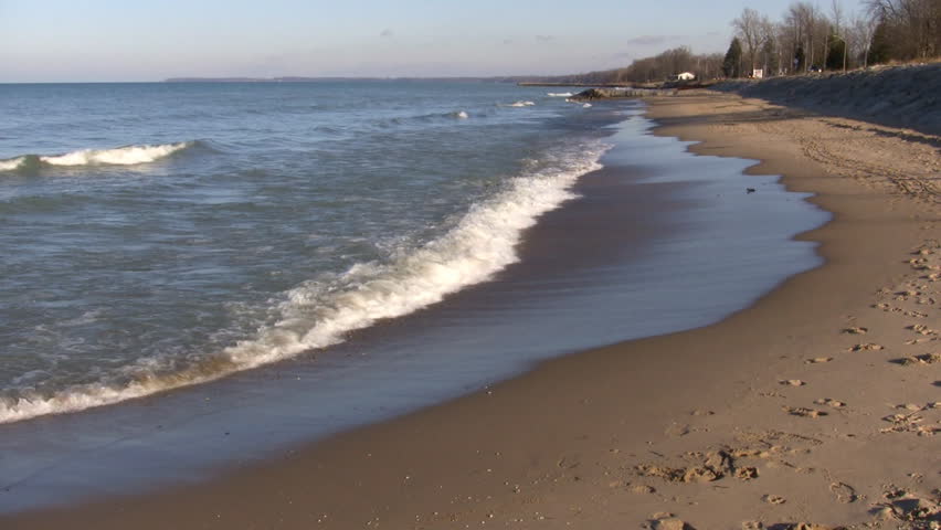 Waves gently hit the beach, then recede as the sunlight illuminates the surf and the blue sky reflects off of the wet sand. (High Definition)