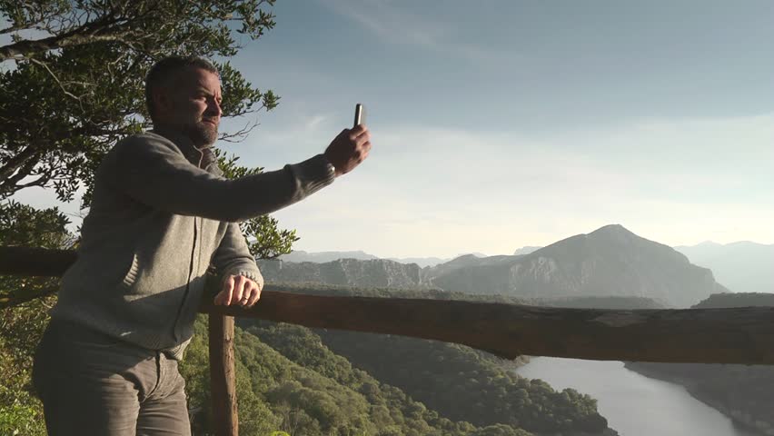 Happy adult man smiling while taking souvenir picture of sunset and natural landscape with camera phone or smartphone. Copy space