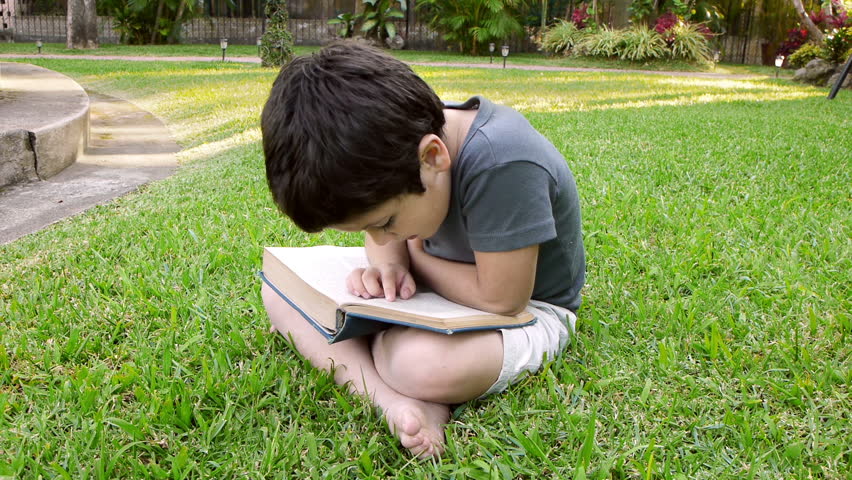 Boy Reading Book (HD). Six year old boy; Hispanic origin flipping the pages of a book while reading to himself sitting on a grassy patch.