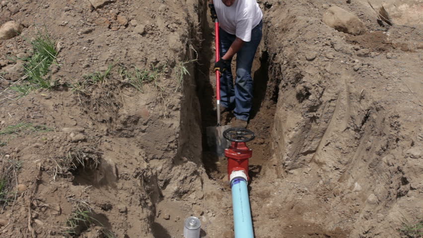 Worker repairs large irrigation water pipe valve in a deep trench