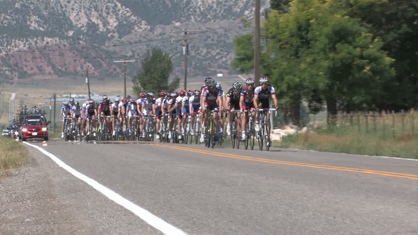 Tour of Utah bicycle race. Coming into a small town of Chester in Sanpete County. Main group close. Group of riders at high speed. Motorcycle with judges and support cars following. 
