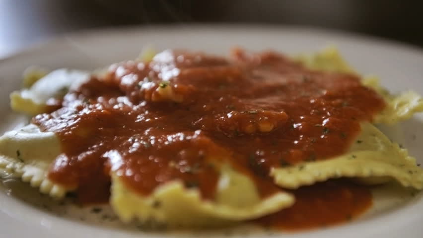Ravioli with tomato sauce in a white plate. Grated parmesan cheese falling from the top and melting on the pasta. 
