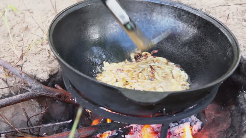 Man mixes broiled meal by colander in pot on campfire