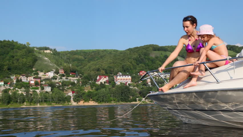 mother and daughter in bathing suits sit on brink of boat on river and talk