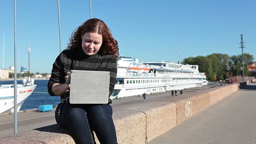 Young beautiful Caucasian woman with tablet computer sitting on city embankment and waving by hand somebody