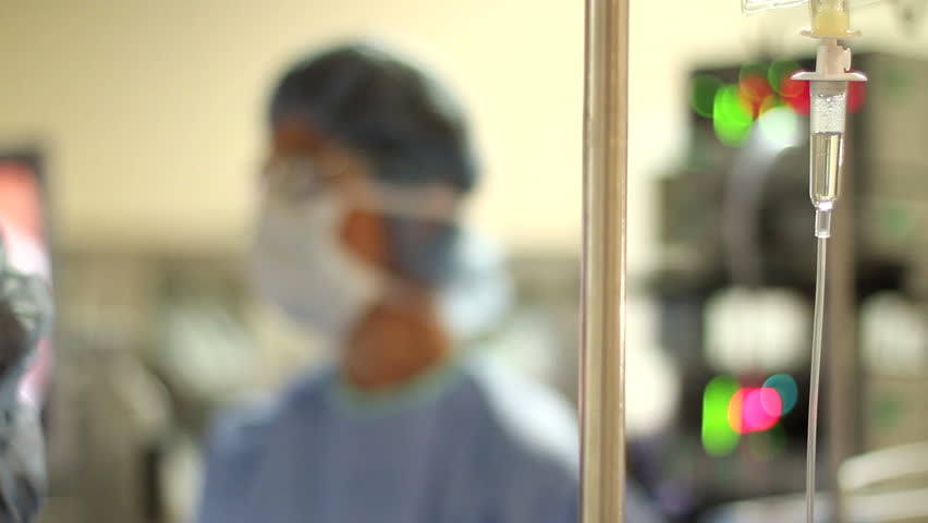Dolly shot of an IV dripping in focus with a surgeon and medical lights & monitors in the background.