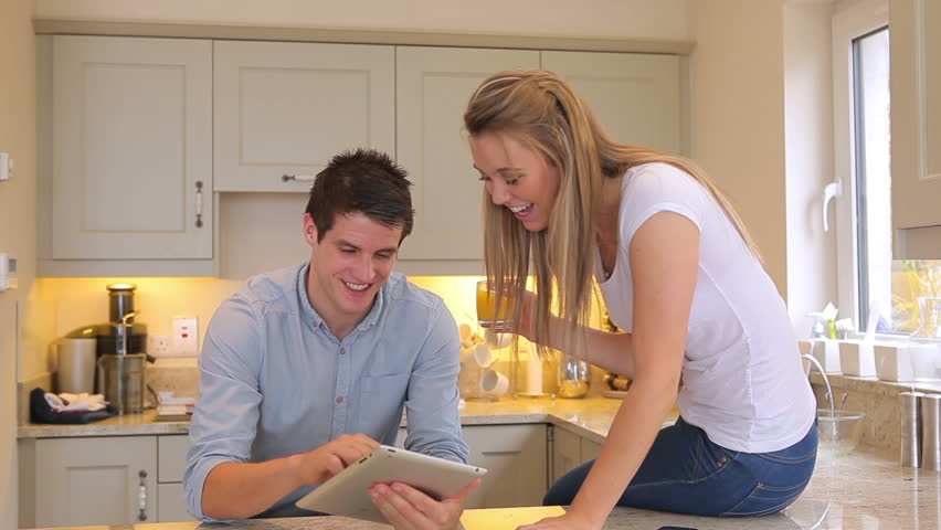 Man showing tablet pc to wife sitting on counter drinking juice in kitche
