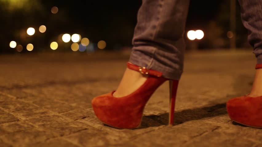 Girl in jeans and red shoes, seen from the side and with close-up on the shoes, pacing along a street at night. Her shadow and the city lights are visible in the background.