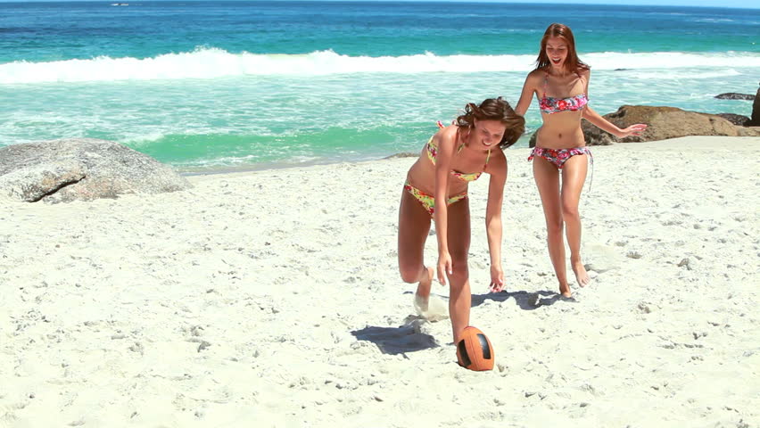 A pair of women in bikinis running towards a rugby ball at the beach