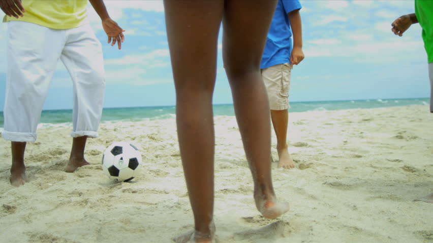 Diverse sons and daughter playing soccer with happy parent on beach
