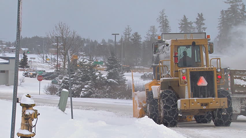 HOMER, AK CIRCA 2012: Snow blower on an excavator paired with a dump truck to clean plow piles on snowy city street.