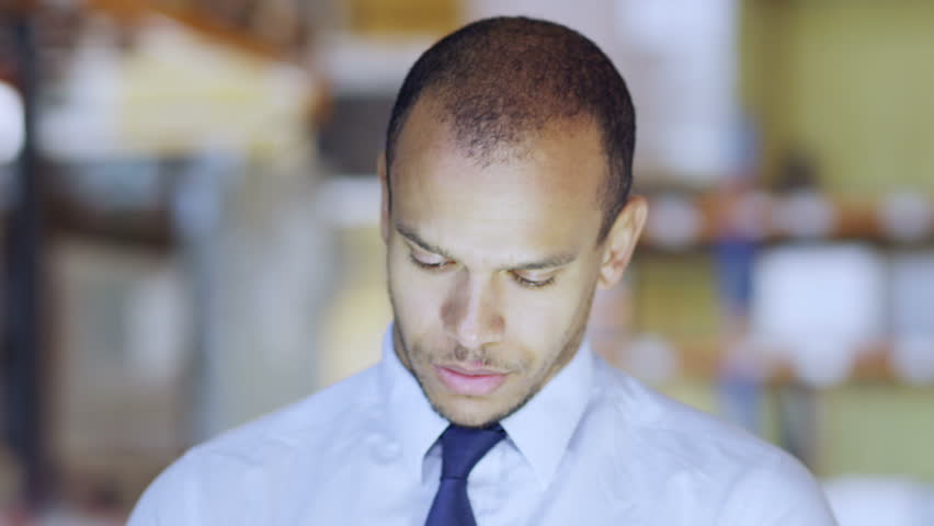 A young man working in a warehouse, dressed in a shirt and tie is working on a digital tablet and checking stock levels. Other employees can be seen working in the background