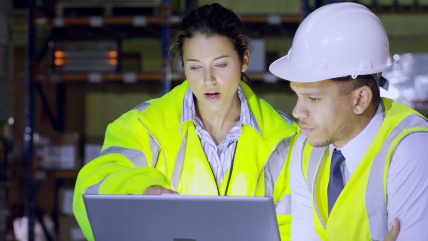 One male and one female warehouse manager are discussing their business and looking at a laptop computer. In slow motion.