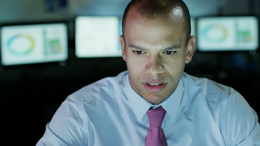 A young businessman is working late at night, with a bank of computer screens behind him. He looks concerned, but then he smiles at something he sees on the computer screen in front of him