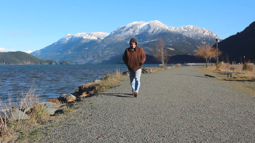 A man walks on a dike or footpath toward camera with a snow capped mountain in the background/Snow Capped Mountain and Footpath/Man walks toward camera with mountain in the background. 