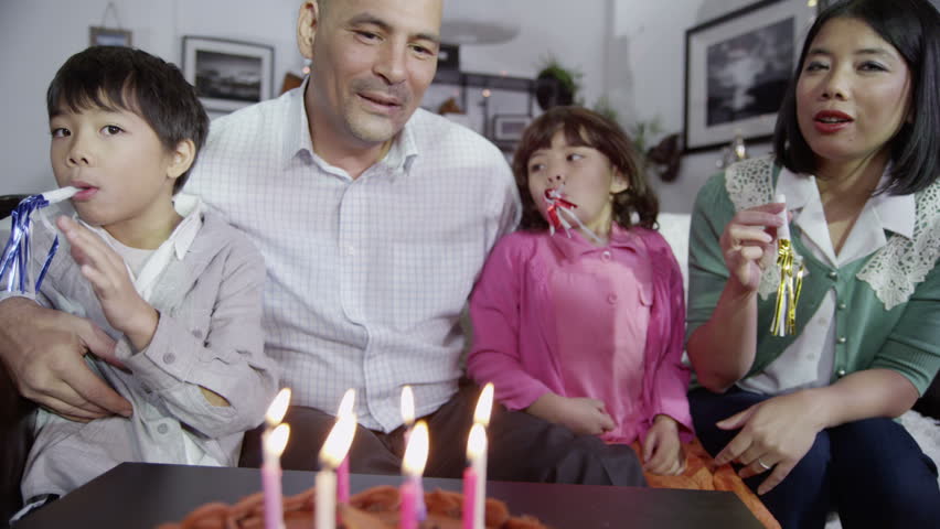 Family at home enjoy a Birthday celebration with party blowers and father blows out the candles on the cake. Interior shot in slow motion.