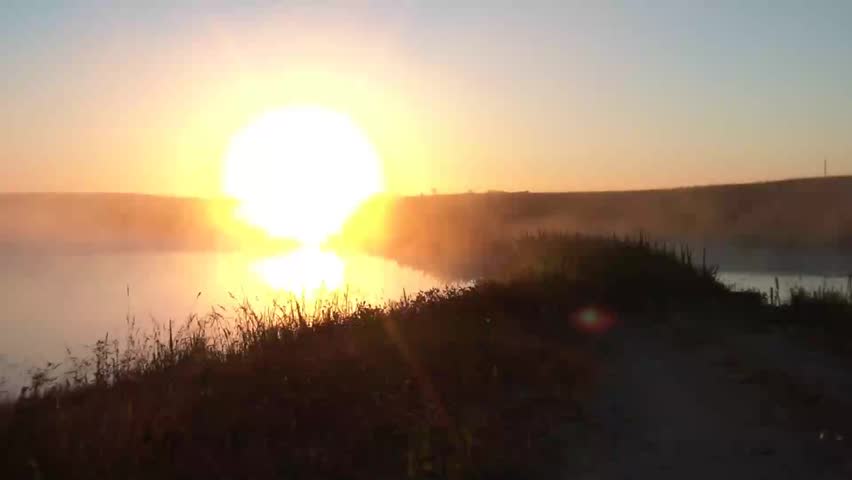 Fog drifts over tranquil lake at sunrise and land with sun burning through, yielding a dreamy diffused look overall.