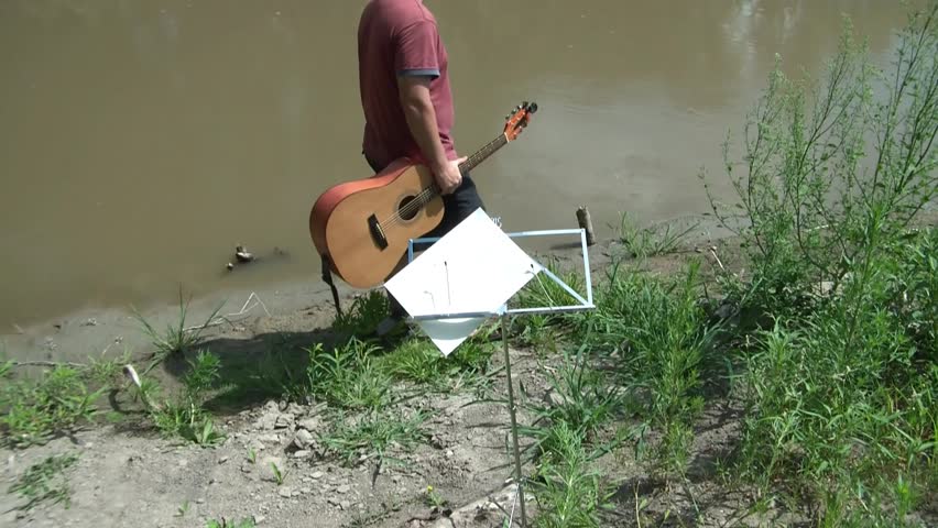Man walks edge of river with acoustic guitar seeking inspiration for songwriting, music note stand in foreground.