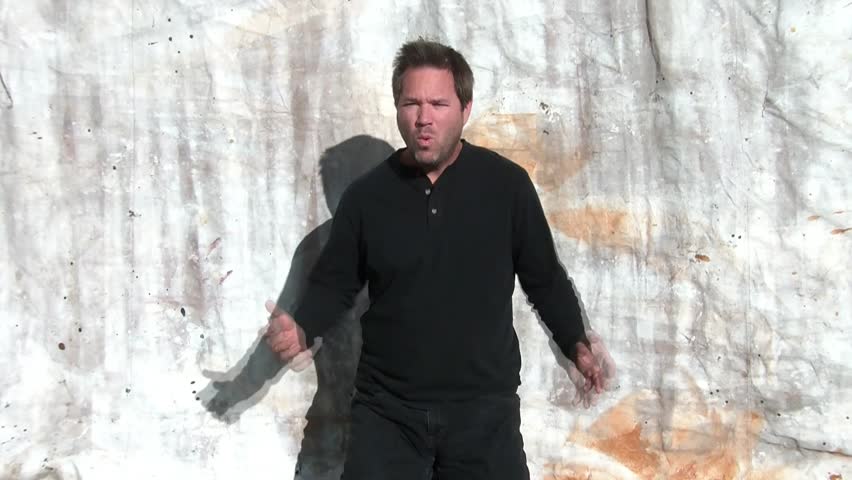 Man spreads arms wide apart and begins mouthing what with attitude, in sunlight, subject wearing black with canvas backdrop.